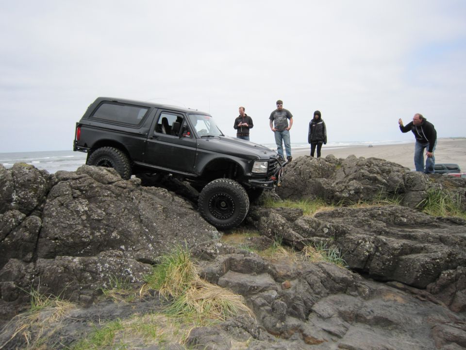 Black Ford Bronco climbing over rocks on a beach with people watching