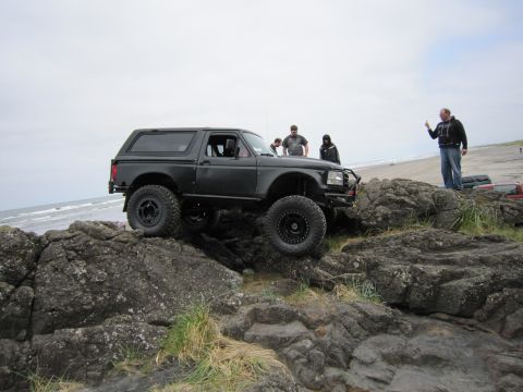 1996 Ford Bronco - A black Ford Bronco navigates a rocky terrain near the ocean with several people observing.