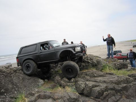 1996 Ford Bronco - A black 1996 Ford Bronco is shown off-roading over rocks on a beach, with several people watching.