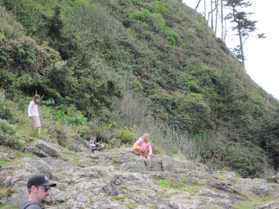 Kids on rocks near a vegetated hill. One girl in pink sits on a rock, another girl stands nearby. A boy is further up the slope