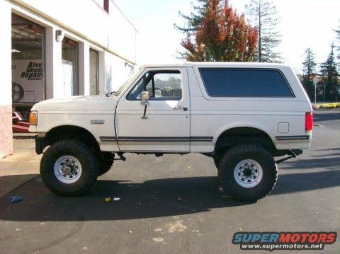 1994 Ford Bronco - Side view of a lifted white Ford Bronco parked outside a repair shop.