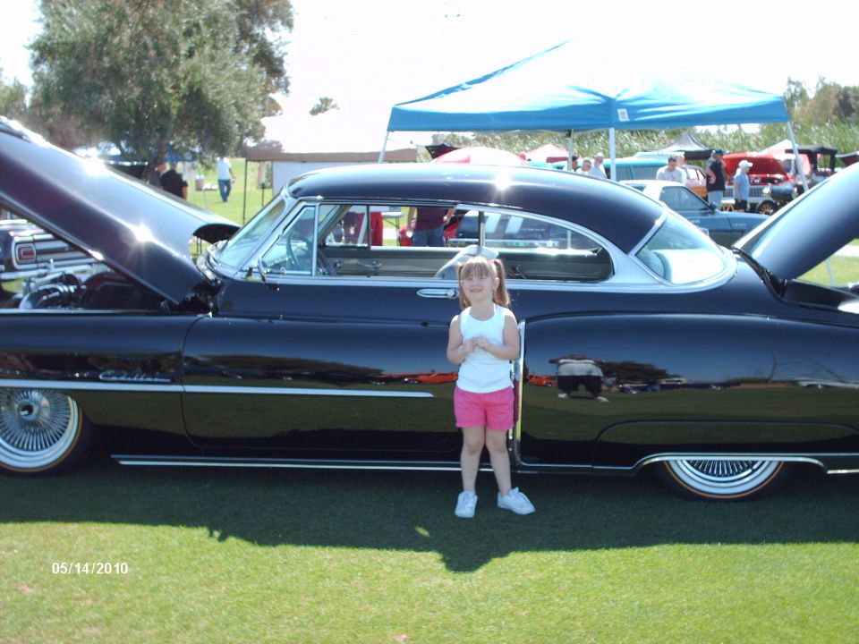 Black coupe with hood open, girl standing beside it