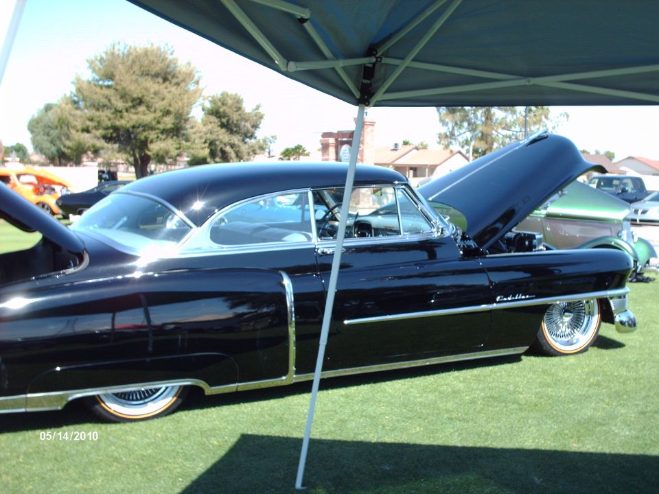 Black 1950s Cadillac with hood open, parked on grass under a canopy. Another car is visible behind it