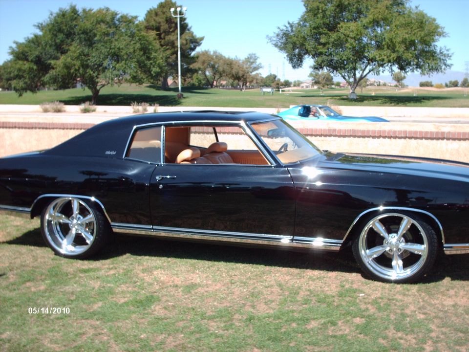 Black coupe with tan interior and chrome wheels parked on grass, with another blue car in the background