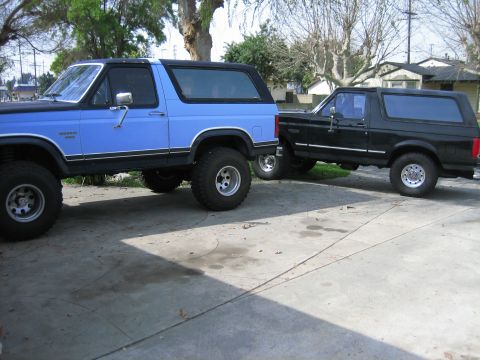 1986 Ford Bronco - Two Ford Broncos, one blue and one black, are parked in a driveway. The blue Bronco is lifted with larger tires.