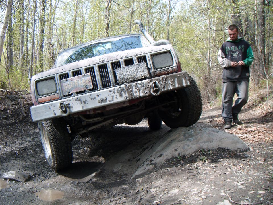 Mud-covered 1997 Jeep Cherokee on a rock obstacle with a man walking nearby
