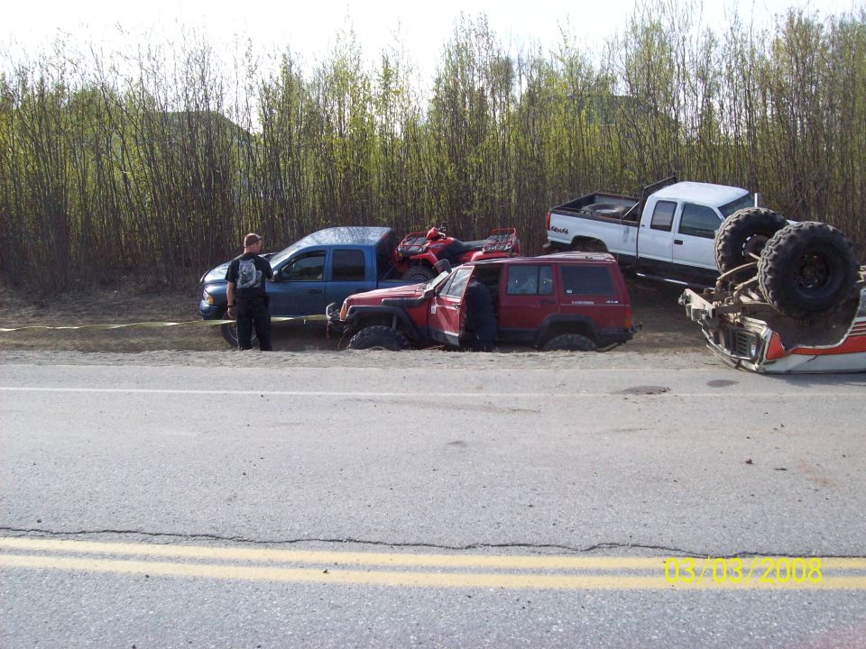 Several off-road vehicles stuck in sand, one overturned on its side near a road