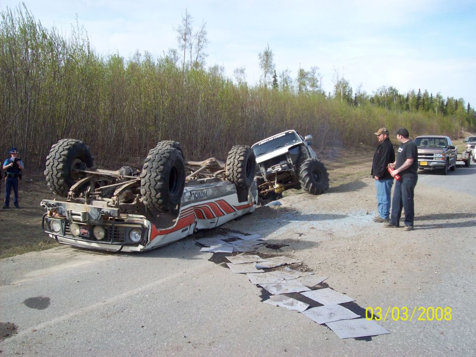 Off-road truck flipped on its side next to a ditch, with two people watching and another truck in the background