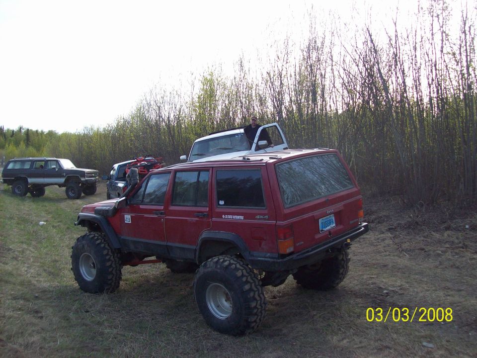 Red Jeep Cherokee XJ with snorkel and oversized tires, rear 3/4 view. Other vehicles visible