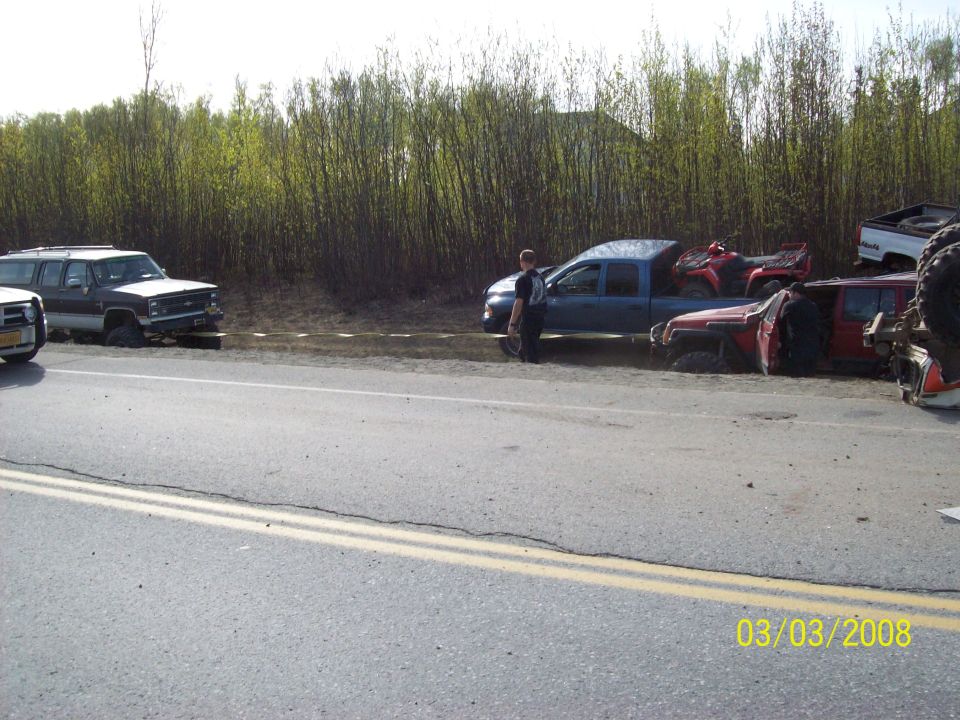 Several vehicles stuck in mud next to a road, including a blue pickup truck with an ATV, a red Jeep, and a dark SUV