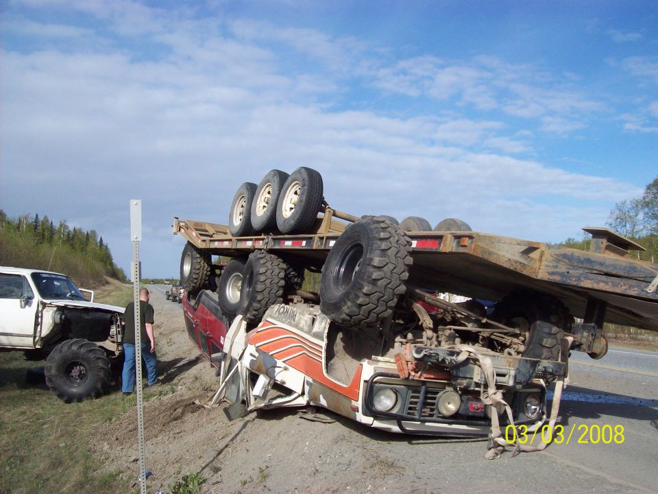Rolled-over truck with trailer on its side, another lifted truck nearby