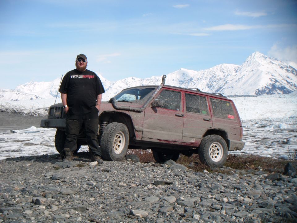 Man standing next to a red 1998 Jeep Cherokee with a snorkel in a rocky, icy landscape