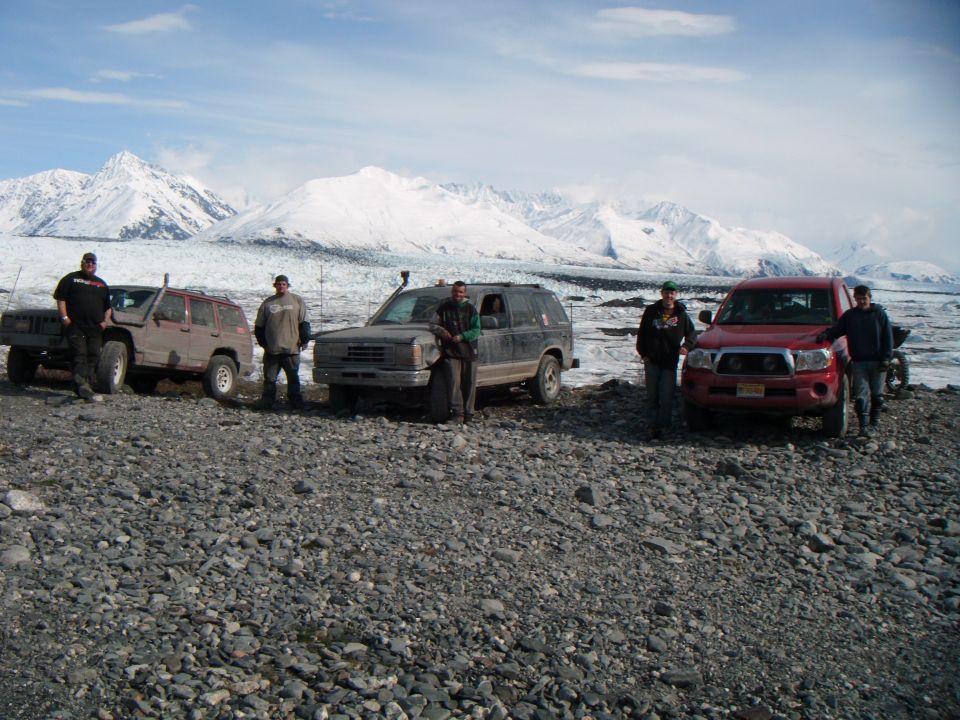 Four people stand with three SUVs and a pickup truck in front of a snowy mountain range and glacier