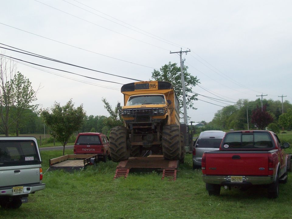 Yellow school bus body on monster truck chassis with huge tires. Other trucks and a trailer are parked nearby