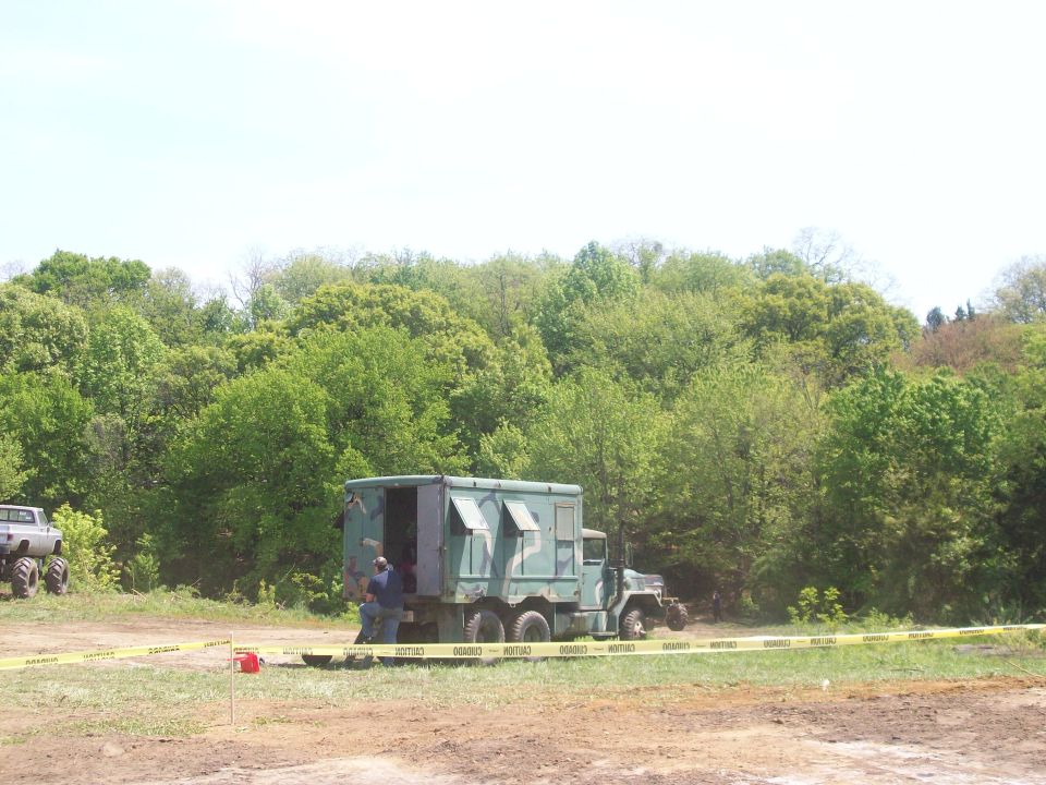 Military truck with a box body parked in a grassy area with trees behind it. Another vehicle with large tires is visible to the left
