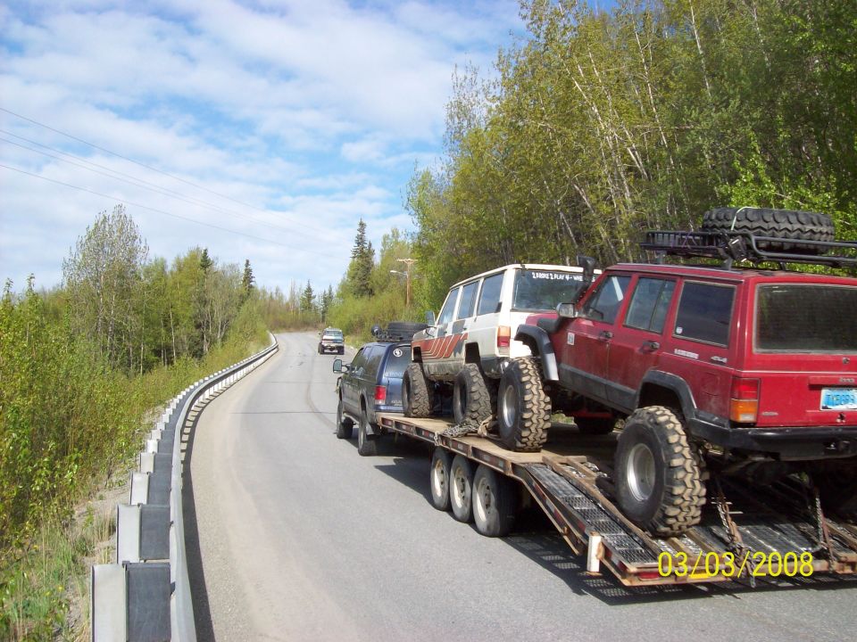 Red Jeep Cherokee and a white Jeep Cherokee on a flatbed trailer being towed on a road