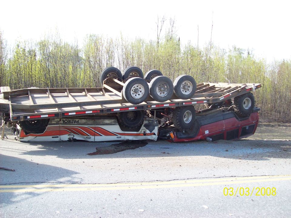 Overturned red SUV with a trailer on top, lying on its side on a road