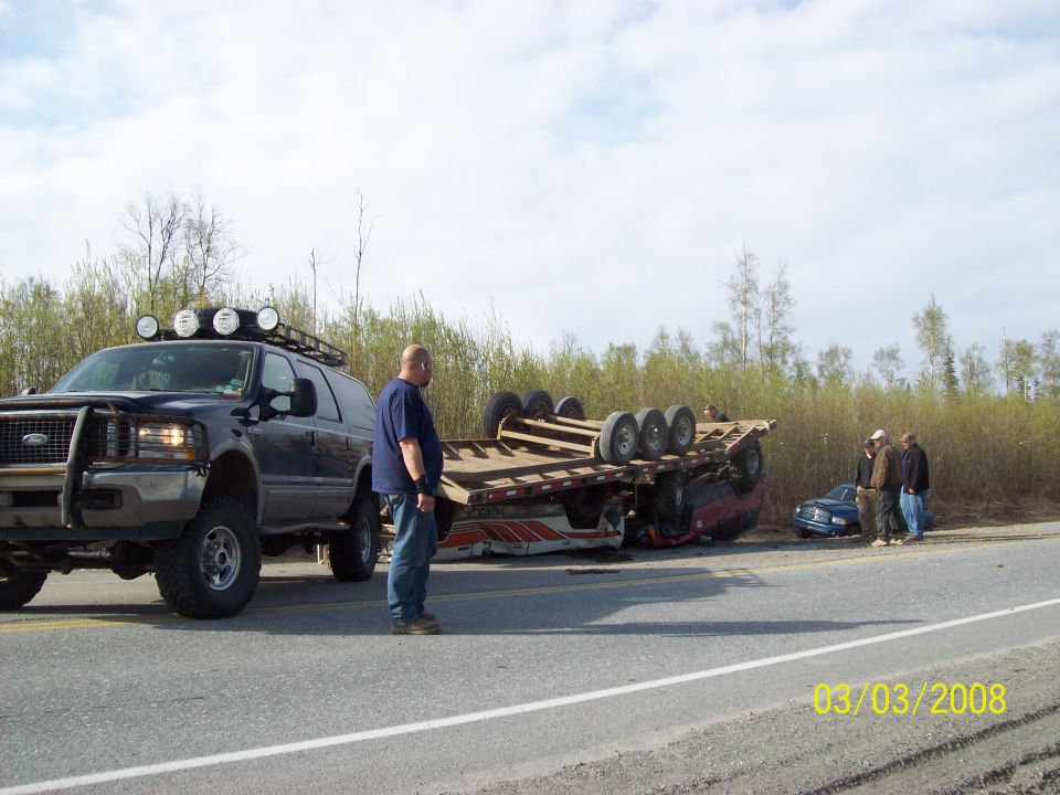 Overturned trailer with car trapped underneath, large SUV on roadside