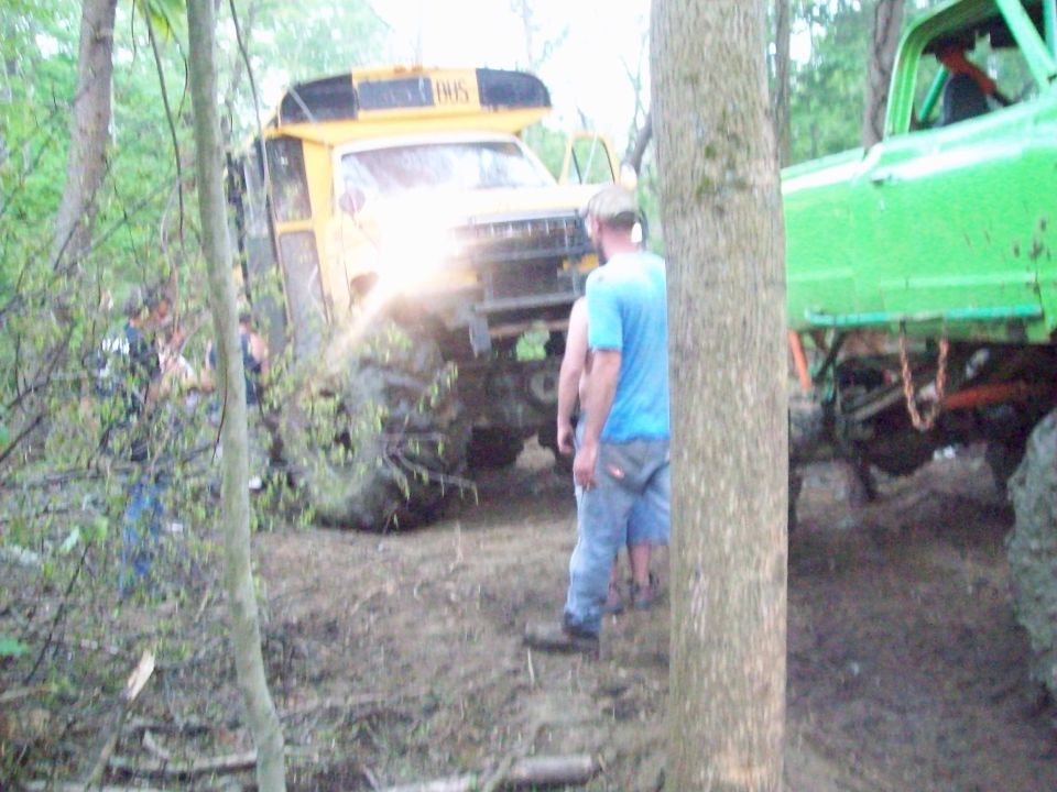 Yellow school bus with "BUS" on top, modified with large tires, stuck in mud with a green truck nearby