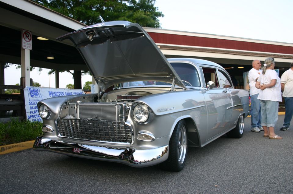 Silver 1955 Chevrolet sedan with hood open, front 3/4 view, chrome grille and bumper, people standing nearby