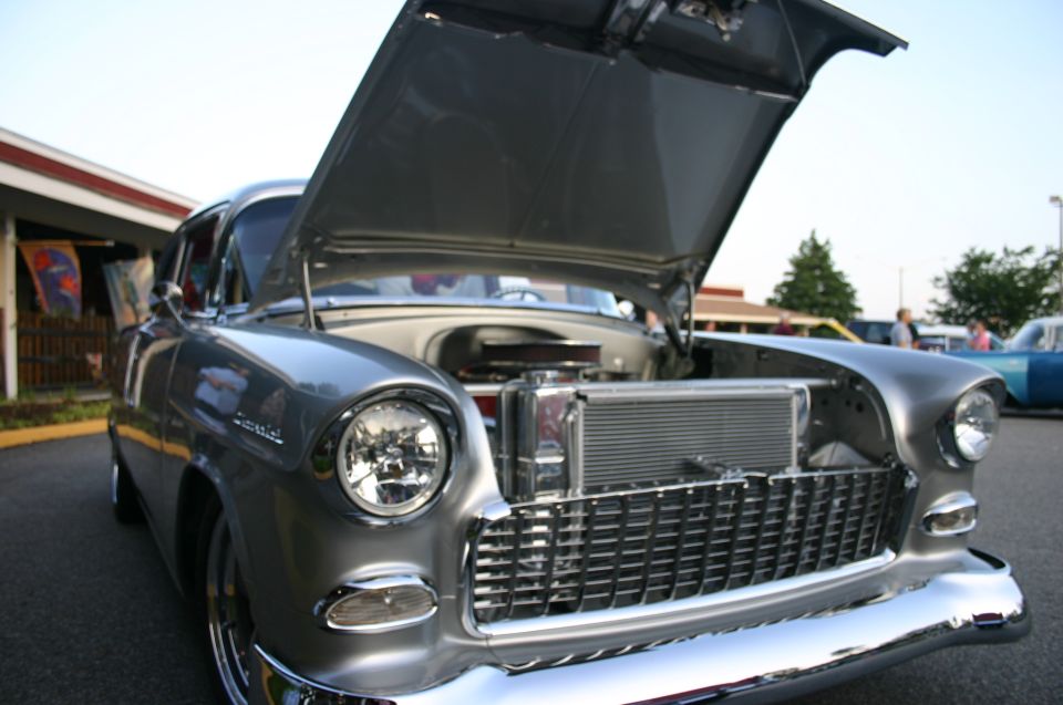 Silver 1955 Chevrolet Bel Air with hood open, showing engine and radiator. Another car is visible in the background