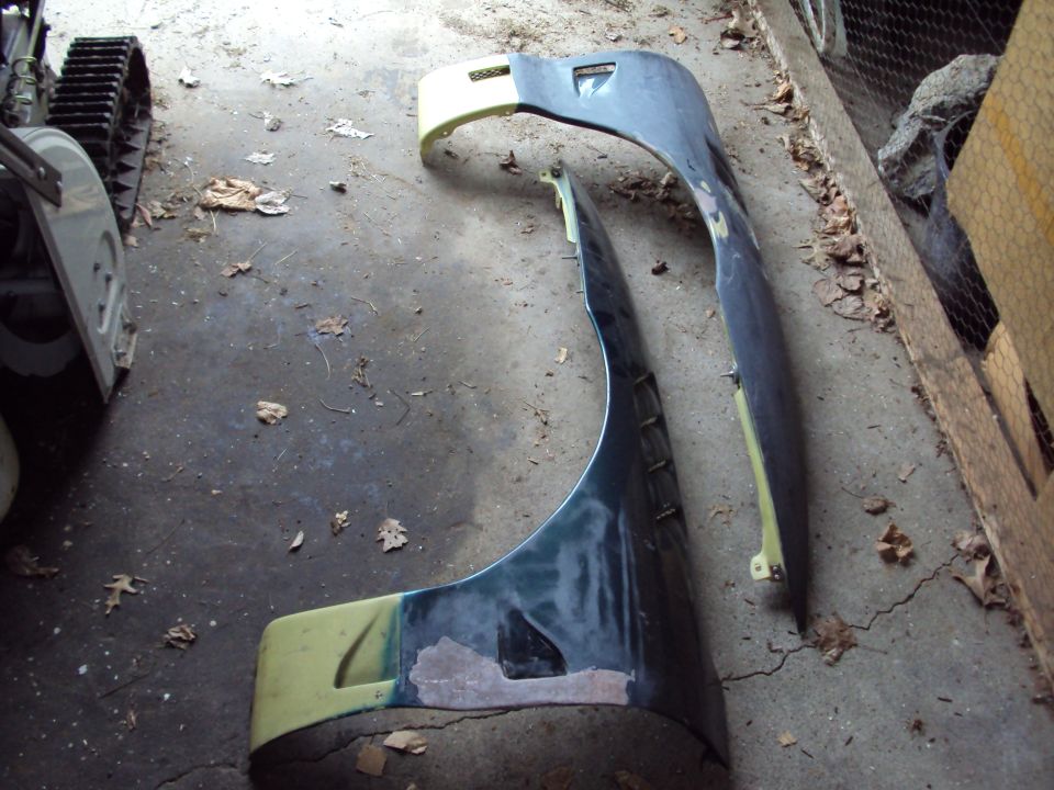 Two car fenders, one dark green and one dark blue, lying on a concrete floor with scattered leaves