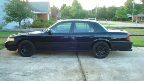 2004 Ford Crown Victoria - A black sedan with black wheels is parked on a concrete driveway in front of a residential house.