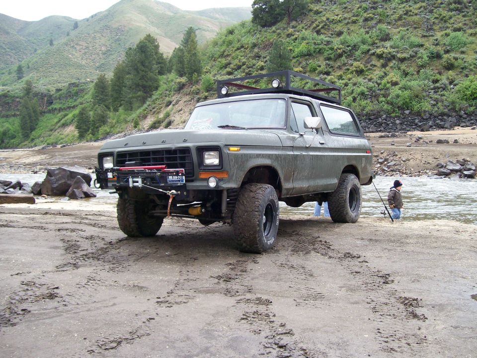 Olive green 1978 Ford Bronco with roof rack parked on a sandy riverbank