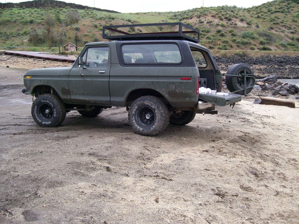 Olive green 1978 Ford Bronco with a roof rack and spare tire on the back, parked on a sandy beach