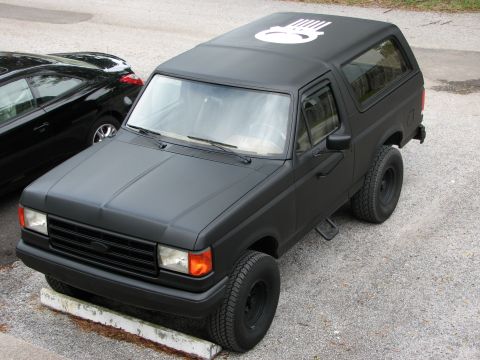 1987 Ford Bronco - This matte black SUV features a distinctive skull graphic on its roof and appears to be a second-generation Ford Bronco.