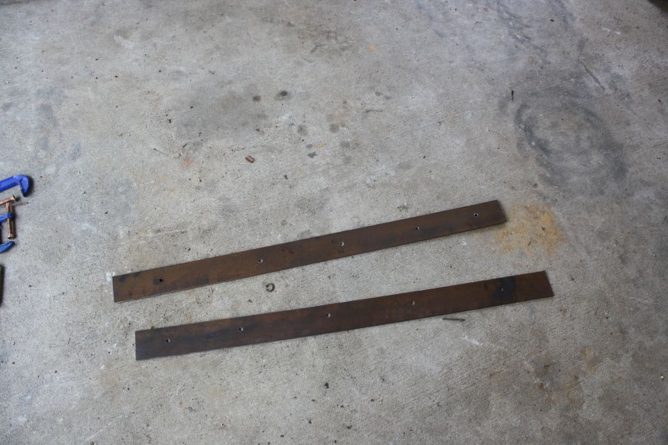 Two rusty metal strips with evenly spaced holes, lying on a concrete floor. A blue clamp is visible on the left