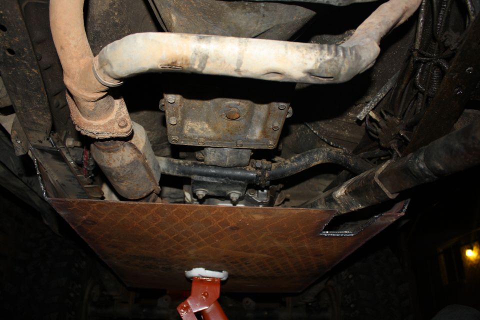 Underbody skid plate and exhaust pipes of a vehicle, viewed from below. A red jack is visible