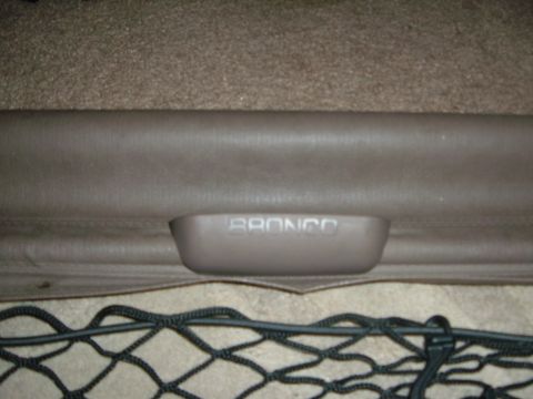 1988 Ford Bronco - Close-up of the tan molded pull handle on a retractable cargo cover, embossed with the "BRONCO" name, set into the vinyl leading edge of th…