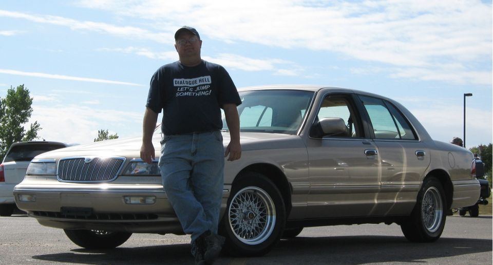Tan 2000 Mercury Grand Marquis, 3/4 front view. Man in graphic tee stands beside it
