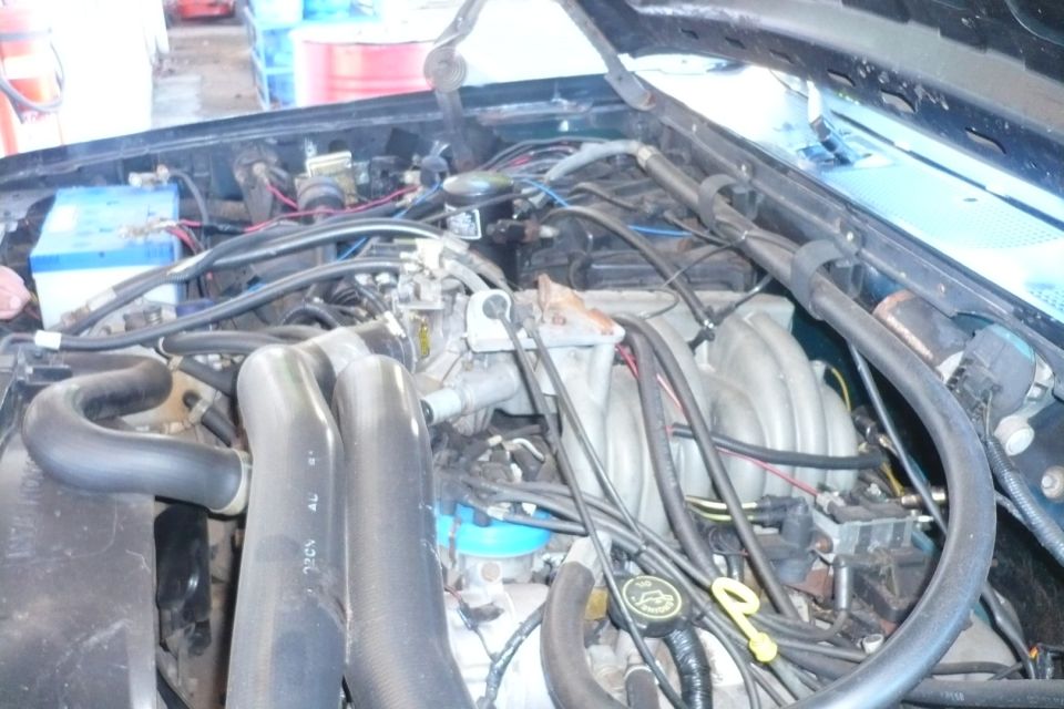 Engine bay with hoses, wires, and engine components visible. A blue battery is on the left