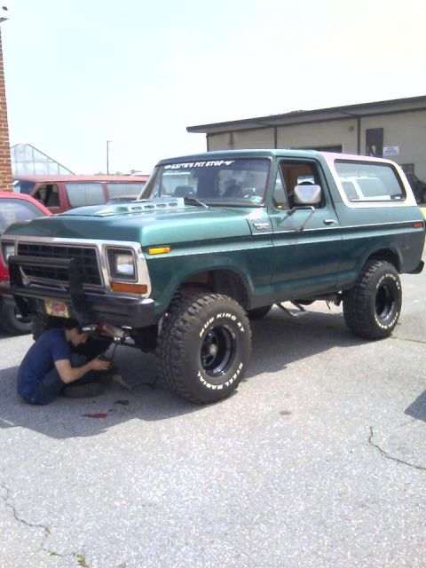 1979 Ford Bronco - A person works on the underside of a green 1979 Ford Bronco with a white removable top and large off-road tires.