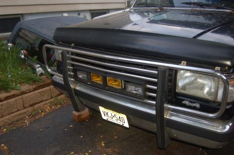 1988 Ford Bronco - The front grille and headlights of a dark blue Ford Bronco are visible, with a chrome bull bar and yellow fog lights.