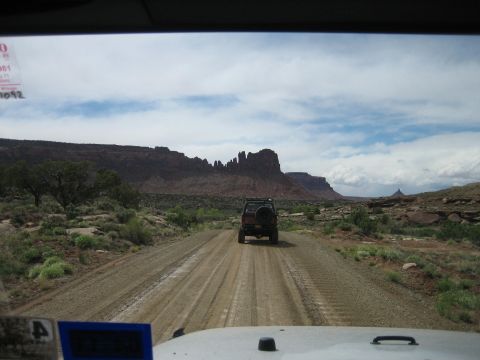 1986 Ford Bronco II - An SUV travels down a dusty road through a desert landscape, with dramatic rock formations under a cloudy sky.