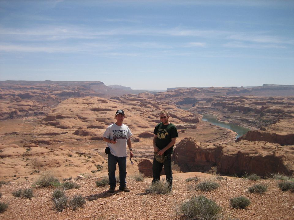 Two men stand on a desert ridge overlooking a vast canyon with a river below. Supermotors.net logo visible