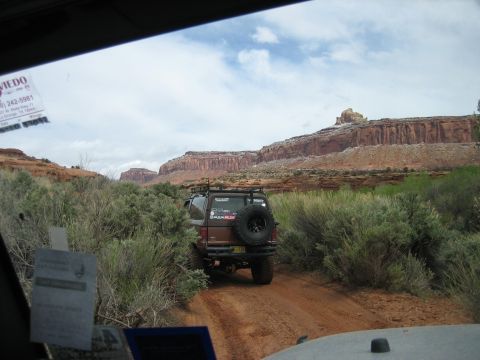 1986 Ford Bronco II - A brown 1986 Ford Bronco II navigates a dusty trail through a scenic canyon landscape, with another vehicle visible ahead.