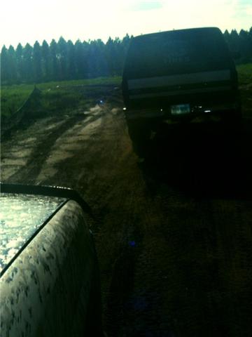 1994 Ford Bronco - Two SUVs navigate a muddy dirt road with trees lining the horizon.