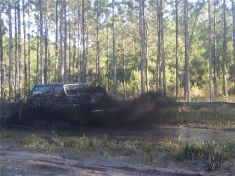 1994 Ford Bronco - An SUV navigates a muddy dirt road, kicking up a large spray of mud as it drives through a wooded area.