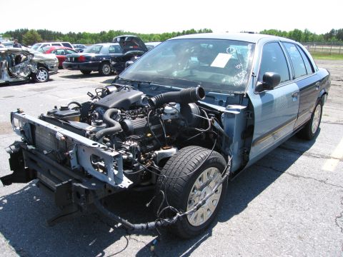 1991 Ford LTD Crown Victoria - A damaged light blue sedan sits in a junkyard with its engine exposed and front end missing.