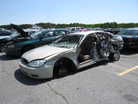 1991 Ford LTD Crown Victoria - A severely damaged sedan sits in a parking lot with its doors ajar and wheels removed.