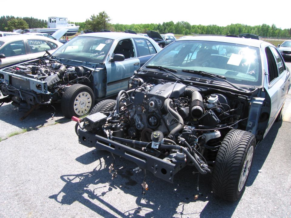 Two stripped 1991 Ford Crown Victoria LTDs in a junkyard, front view, showing exposed engines and missing body panels