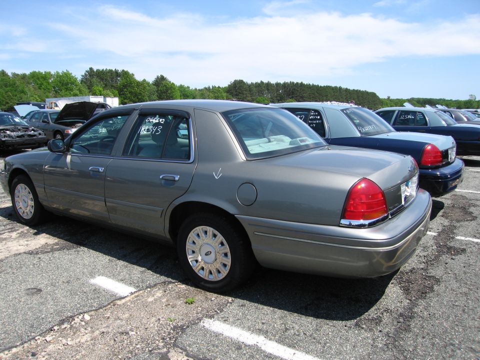Gray Ford Crown Victoria parked in a lot with other cars, rear passenger side view. White markings on rear window