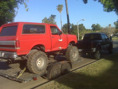 1969 Ford F-250 - A red lifted SUV is transported on a trailer pulled by a black pickup truck under a clear blue sky.