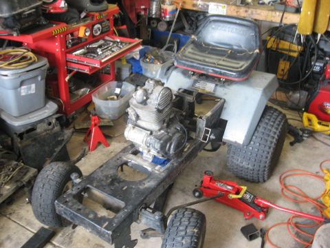 2010 Kawasaki BAYOU 220 - A lawn tractor is in a workshop, partially disassembled with its engine exposed and supported by jacks.