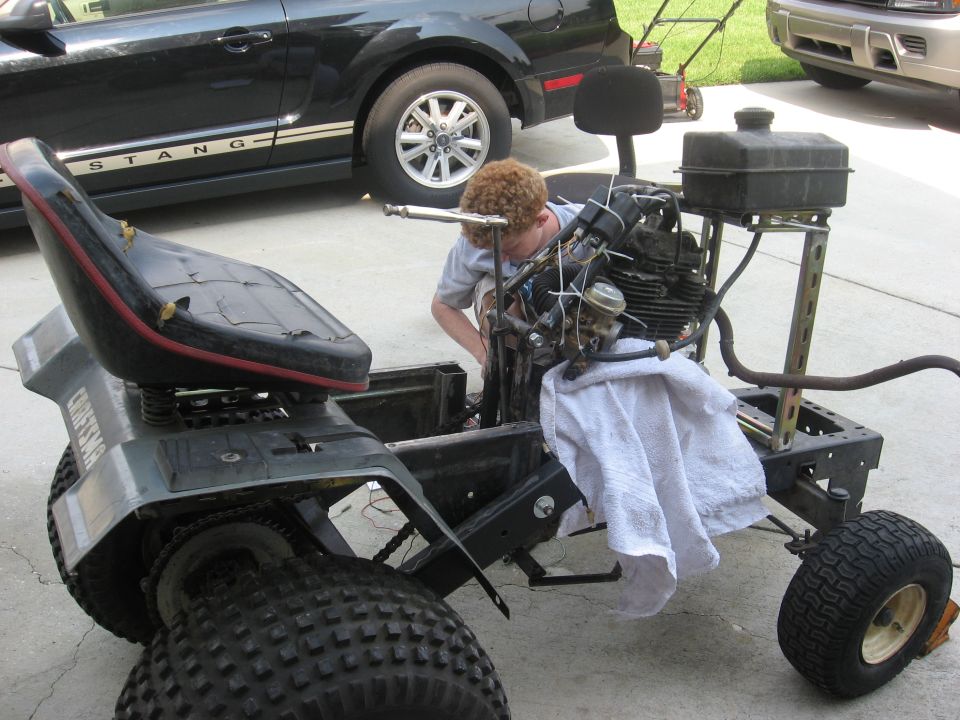 Person working on a Craftsman lawn tractor chassis with engine exposed. Black Ford Mustang in background