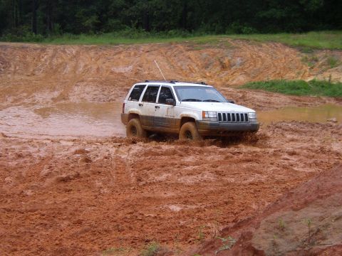1991 Ford Bronco - A white Jeep Grand Cherokee navigates a challenging muddy track, kicking up dirt as it goes.